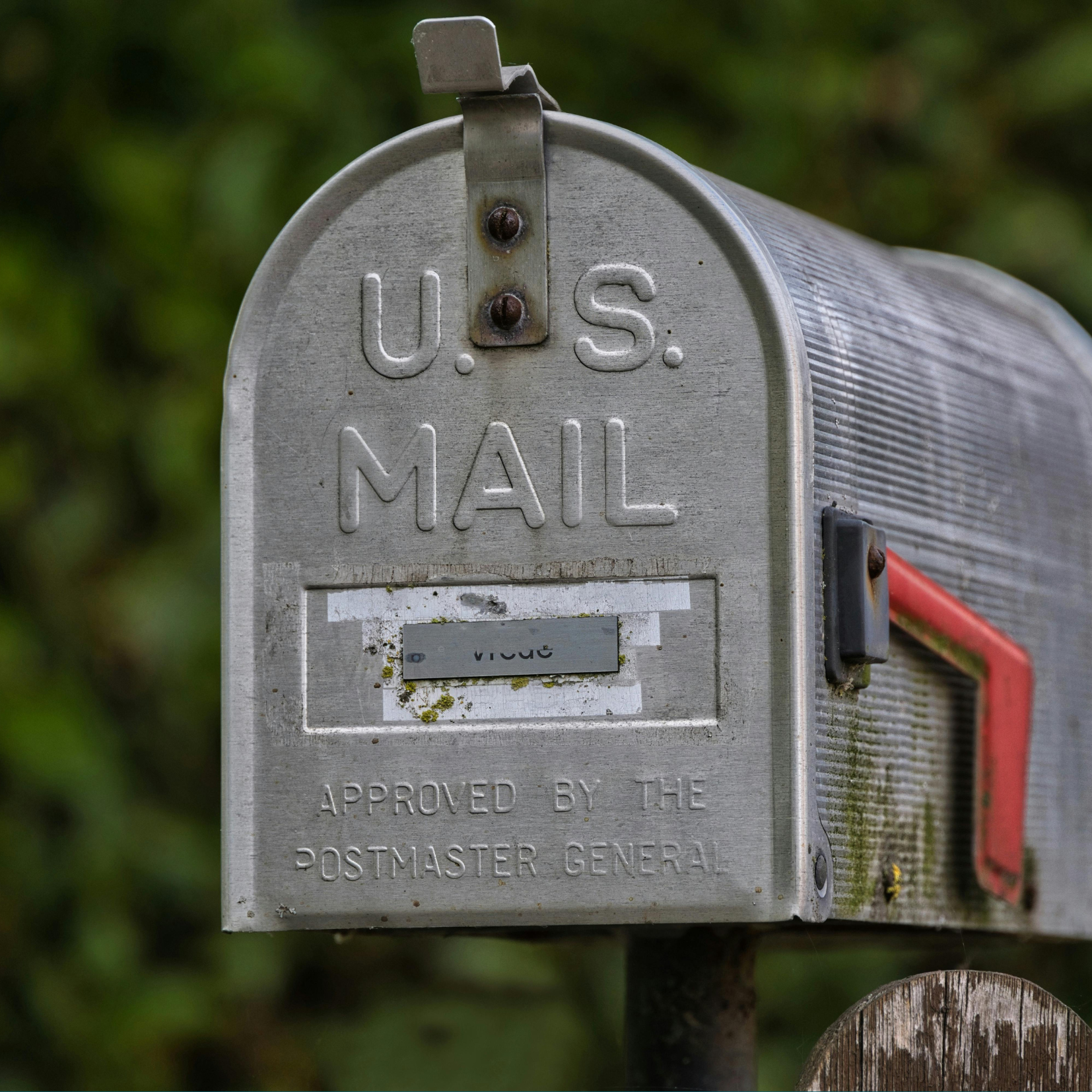 A close-up of a traditional silver U.S. Mail mailbox with a red flag, mounted on a wooden post. The surface shows some wear and moss, and the background is blurred greenery.