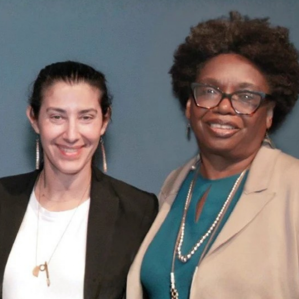 Two women standing side by side and smiling at the camera—one with dark hair tied back in a black jacket, the other with short curly hair, glasses, and a beige blazer—showcasing their dedication to advocating for renters and protecting renters rights.