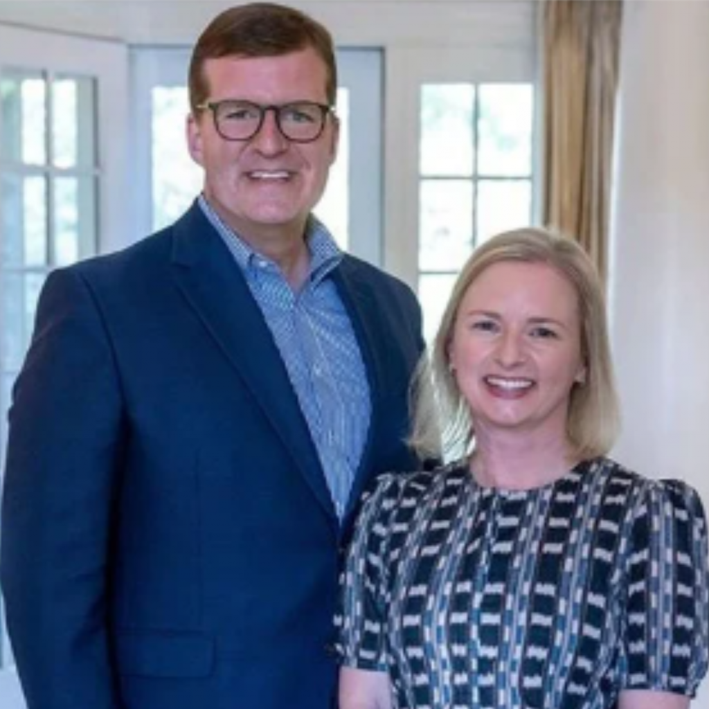A man in a blue suit and glasses stands next to a woman with blonde hair in a patterned dress. Smiling and posing indoors by large windows, they embody giving with purpose through their philanthropic partnership.