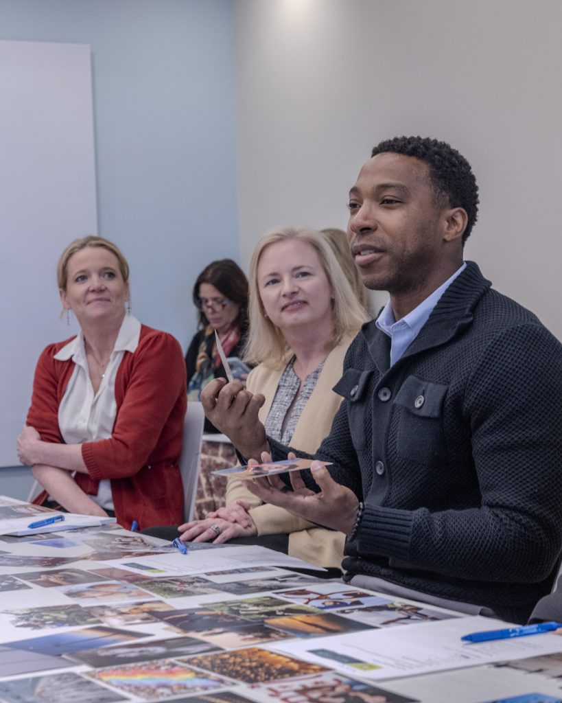 A man speaks and gestures while sitting at a table with papers and photos, surrounded by three women who listen attentively during a philanthropy consulting meeting in a conference room.