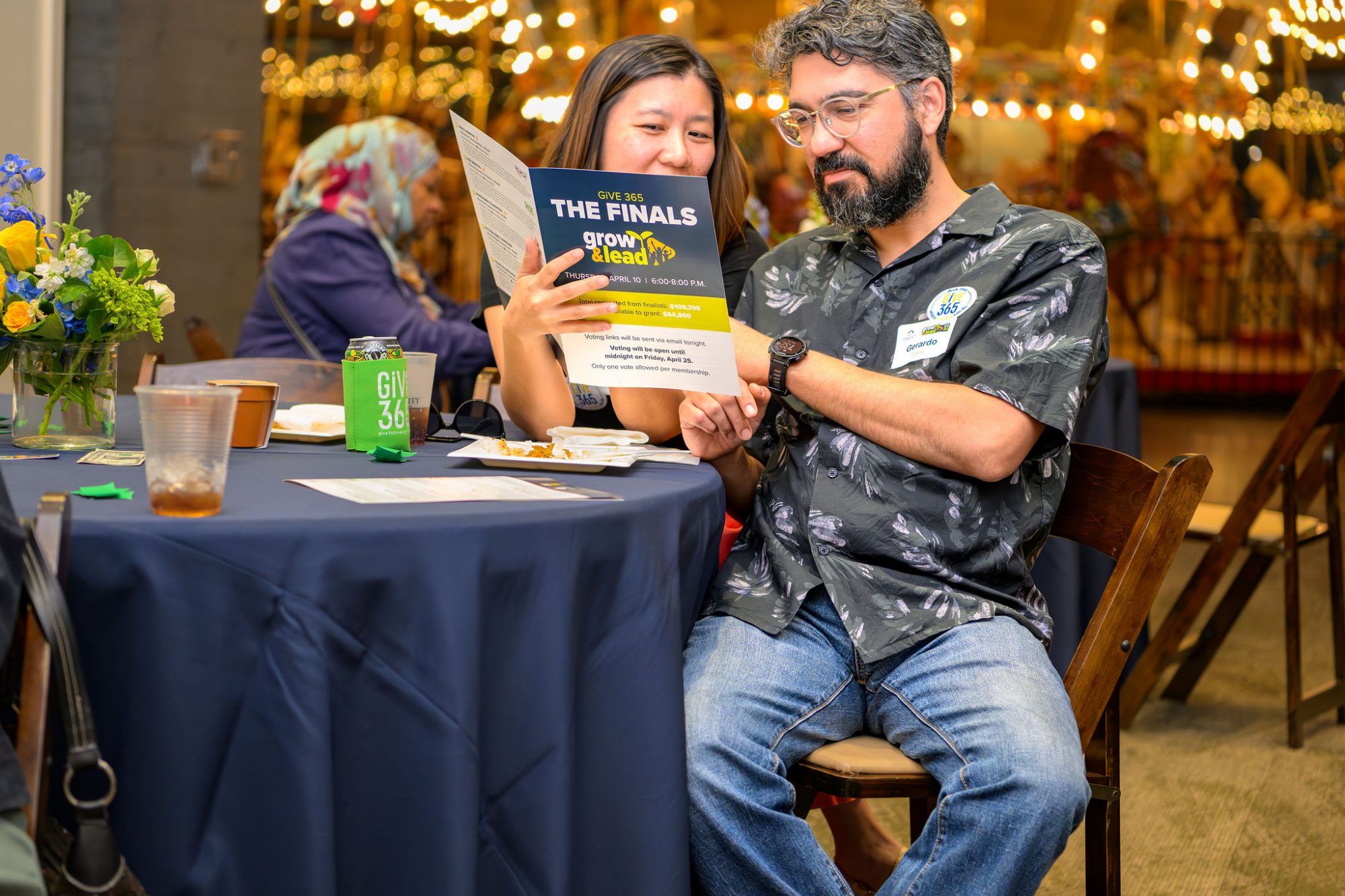 Two people sit at a table, looking at a flyer titled THE FINALS grow to lead. The man wears glasses and a floral shirt; the woman smiles while holding the flyer. A drink can and flowers are on the table. A carousel is in the background.