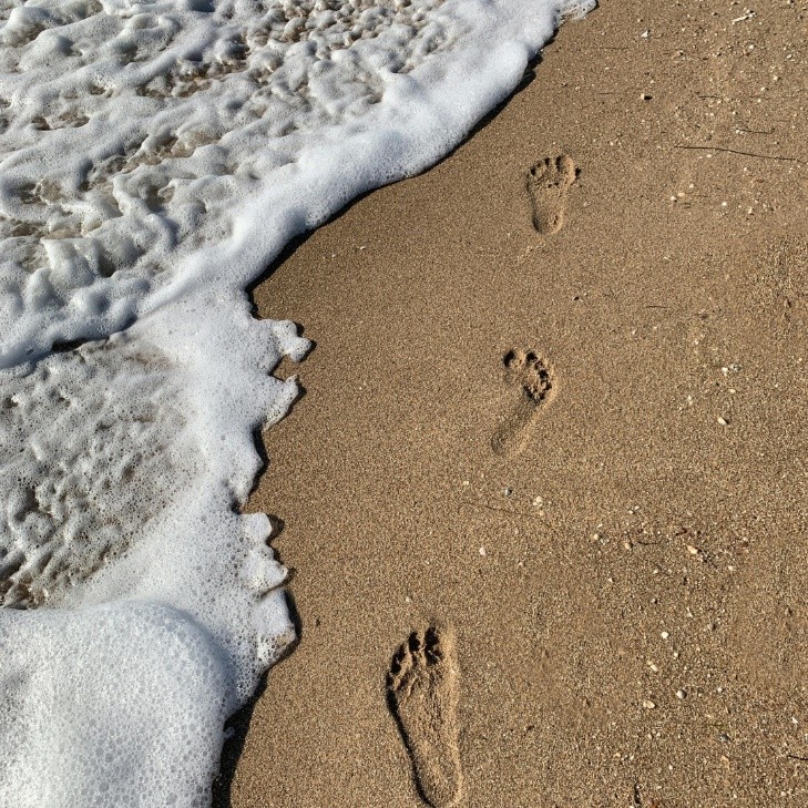 Three footprints in the sand are being approached and partially washed away by gentle ocean waves and white foam, evoking a sense of sudden life changes unfolding along the tranquil shore.