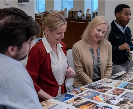 Four people, including Jessica and Sandy Webb, sit around a table covered with various photographs, smiling and discussing the images in a brightly lit office—a warm moment of giving with purpose.