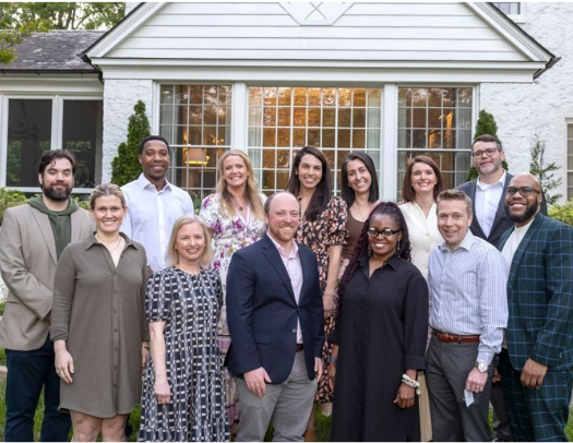 A group of thirteen adults, including Jessica and Sandy Webb, dressed in business casual attire, stand together and smile outside a white house with large windows and greenery, celebrating their philanthropic partnership.