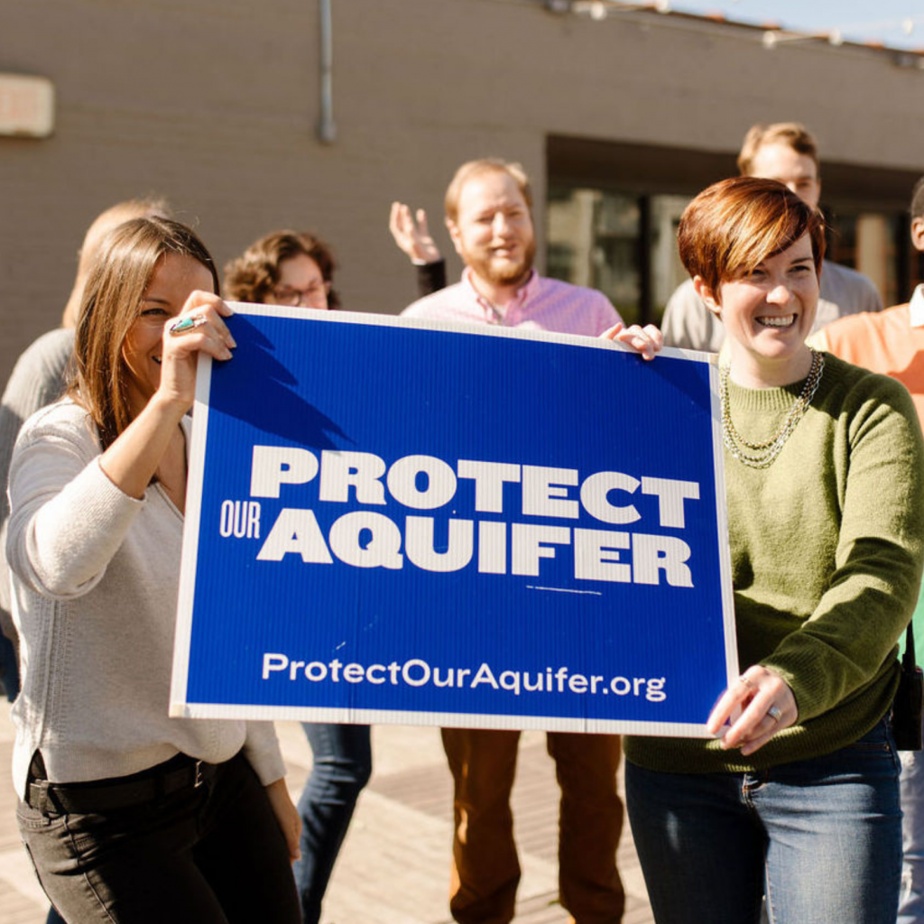 A group of smiling people stand outdoors holding a large blue sign that reads PROTECT OUR AQUIFER and ProtectOurAquifer.org. The mood is positive and energetic.