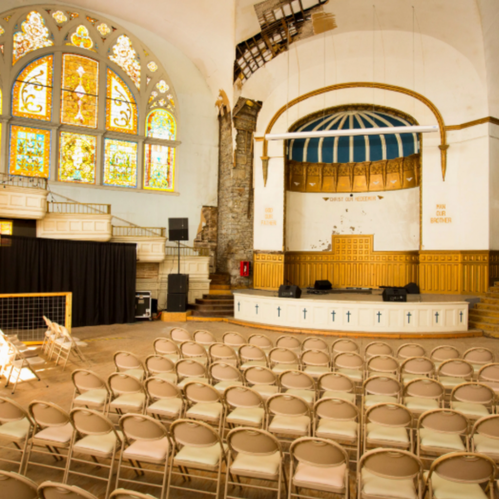 Rows of beige chairs face a stage in a bright church hall with large stained glass windows, an arched ceiling, and gold and white architectural details. Audio equipment is set up near the stage.