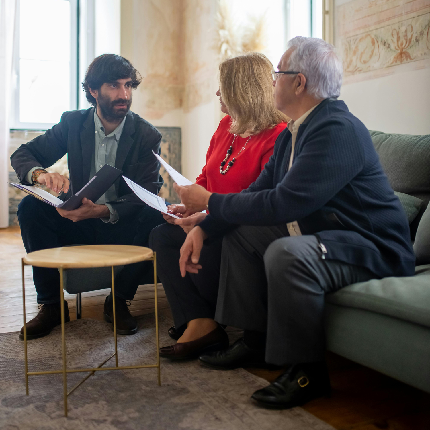 Three people sit together indoors, dressed in business attire, having a discussion while holding papers. One man points to a document, engaging with the woman and another man seated on a couch beside him.