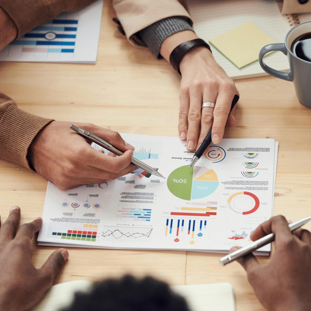 Three people sit at a table, reviewing colorful charts and graphs on a paper. Two hands point at the document with pens, while another hand holds a pen nearby. A coffee mug and notepad are also on the table.
