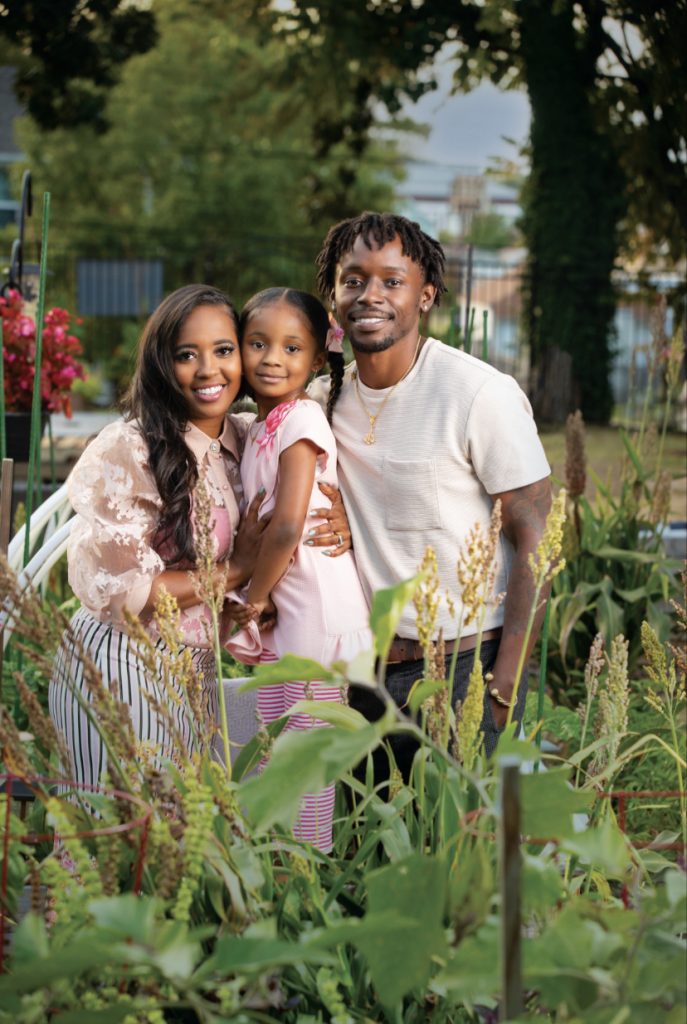 A smiling family—two adults and a young girl—pose together in a lush community garden surrounded by green plants and flowers on a sunny day.