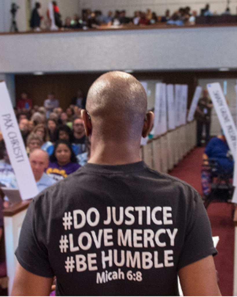 A person stands at the front of a crowded church, seen from behind, wearing a shirt that reads #DO JUSTICE #LOVE MERCY #BE HUMBLE Micah 6:8. Congregants sit in pews with banners along the aisles, celebrating recent grants supporting their mission.