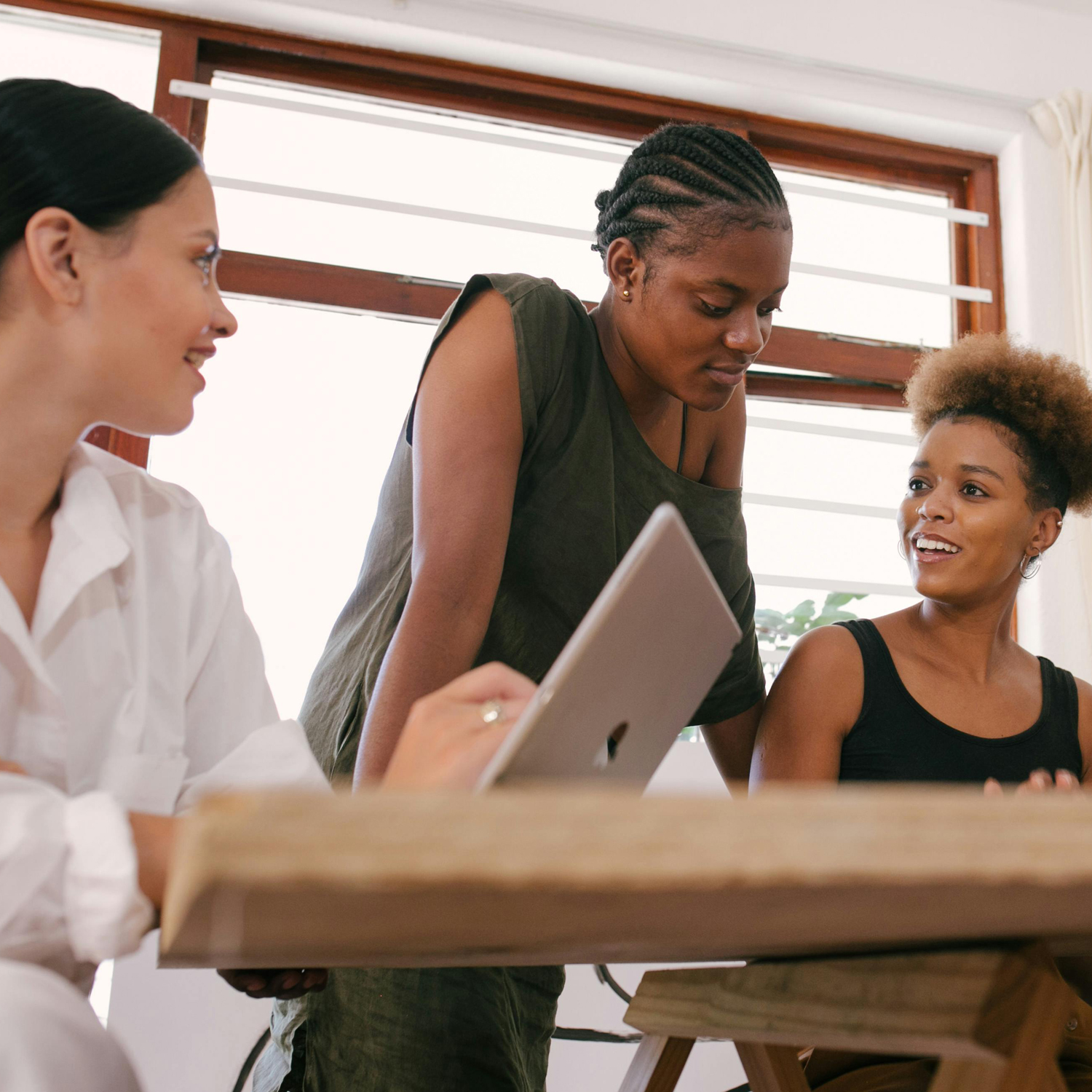 Three women sit and stand around a desk, engaged in conversation. One woman holds a laptop, while the others listen and smile. They are in a brightly lit room with large windows in the background.