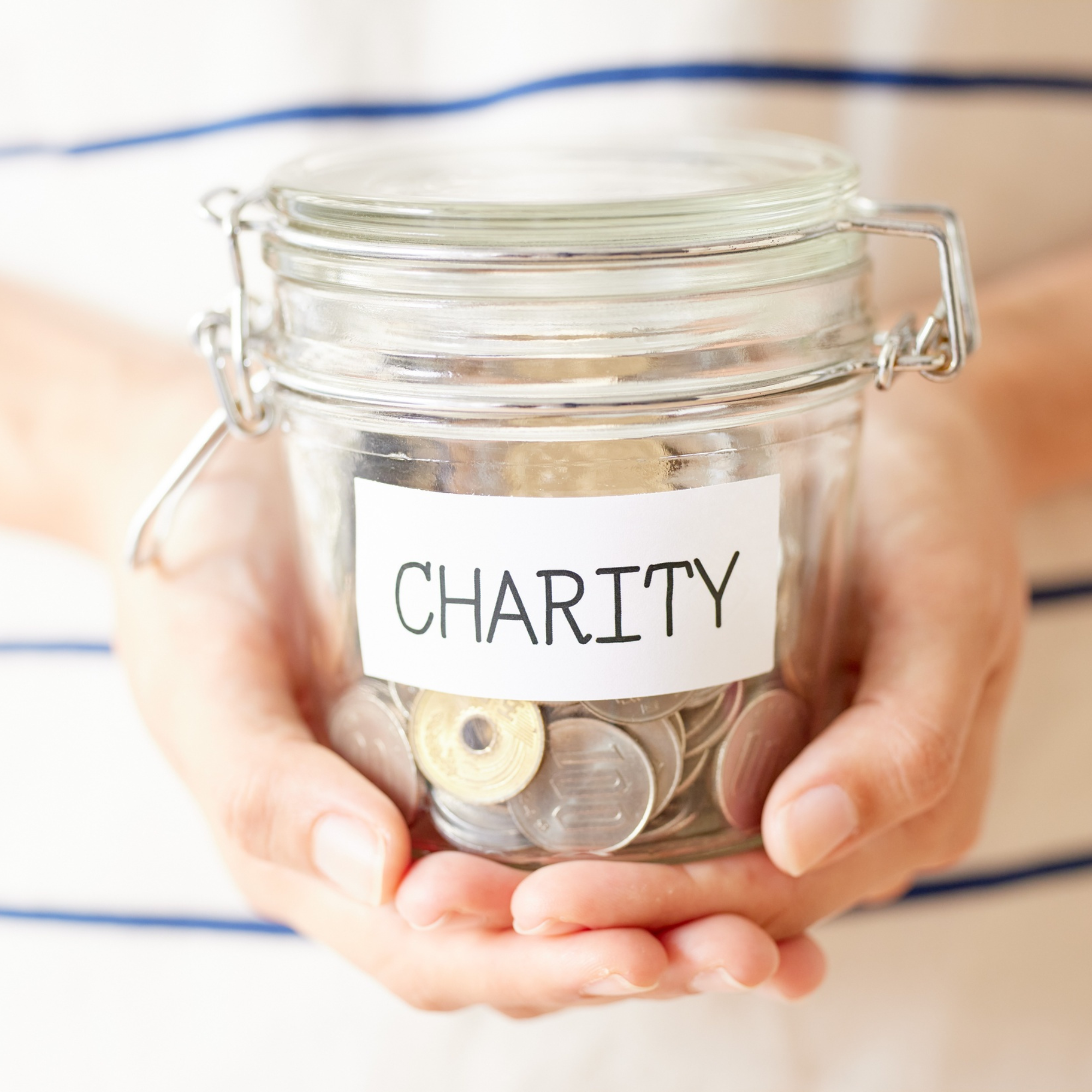 A person holds a glass jar filled with coins. The jar has a white label with the word CHARITY written on it in bold, black letters, symbolizing support for charitable clients through dual strategies of giving and serving charities.