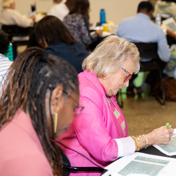Two women sit at a table in a classroom or meeting setting, focused on reading and highlighting documents, with other people working in the background.