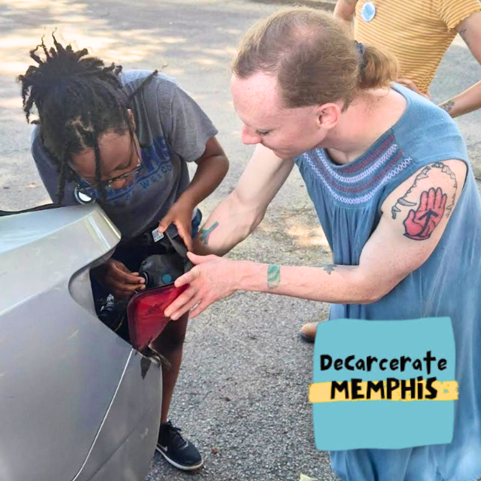 Two people work together to attach a taillight to a silver car—one holds the light while the other uses a tool. A Decarcerate Memphis sticker sits in the corner, sharing stories that help heal communities. Another person stands in the background.