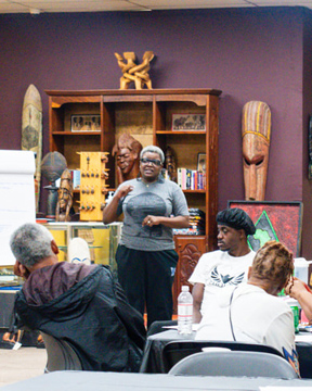 A person stands and speaks to a seated group in a room decorated with African art and masks, fostering healing through sharing stories. A flip chart, wooden bookshelf, and colorful artwork are visible in the background.