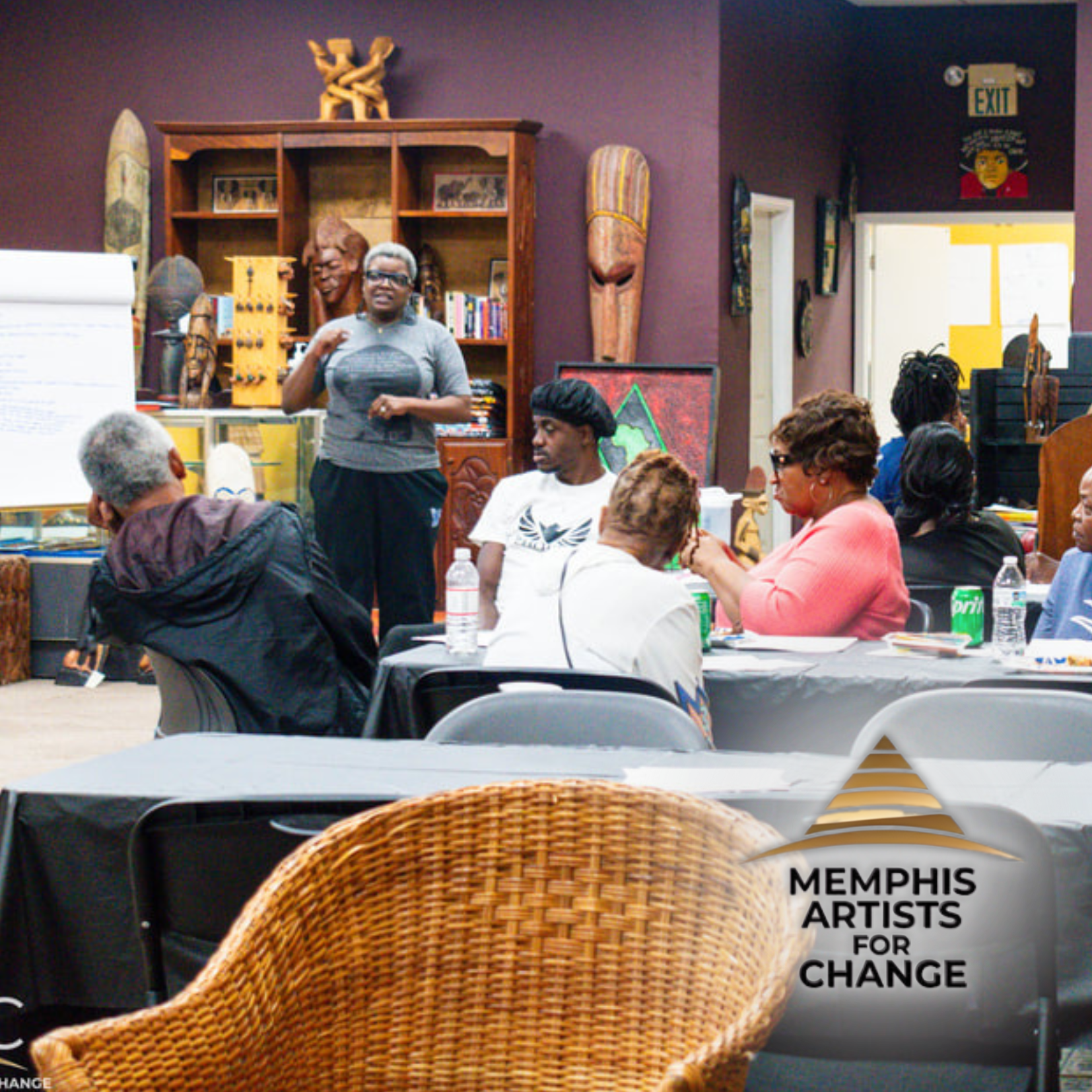 A group of people sit around tables in a room with African art while a person stands and speaks at a flipchart, highlighting youth empowerment. The Memphis Artists for Change logo appears in the foreground.