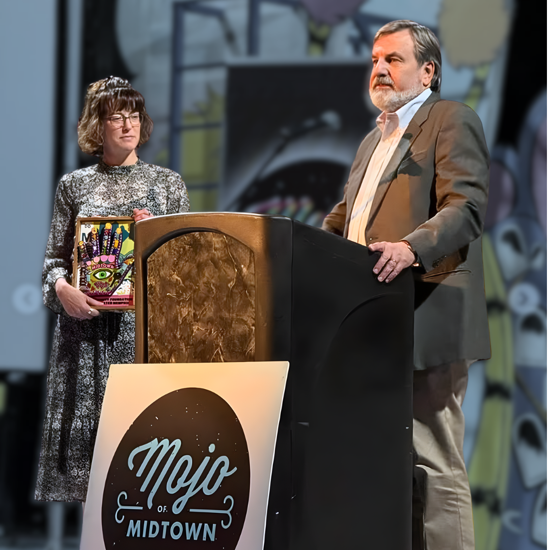 A woman holding a colorful award stands next to a man speaking at a podium with a Mojo of Midtown sign, against a backdrop featuring abstract artwork.