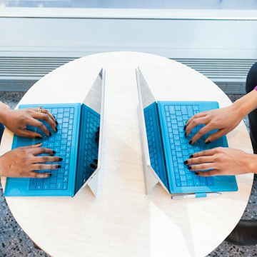 Two people sit across from each other at a round table, engaged in client communication, typing on blue keyboards with their laptops facing each other. Their hands are visible as they discuss transferring a private foundation.