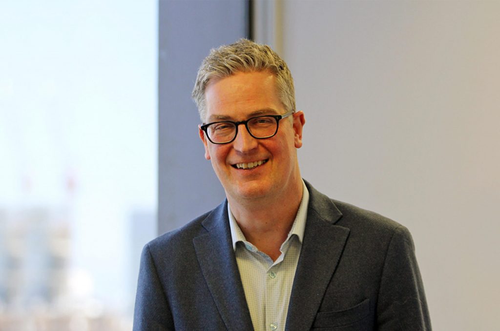 A smiling man with short, gray hair and glasses wearing a blue blazer and light shirt stands indoors near a window, the blurred cityscape outside hinting at the excitement of the upcoming 2026 Annual Meeting.