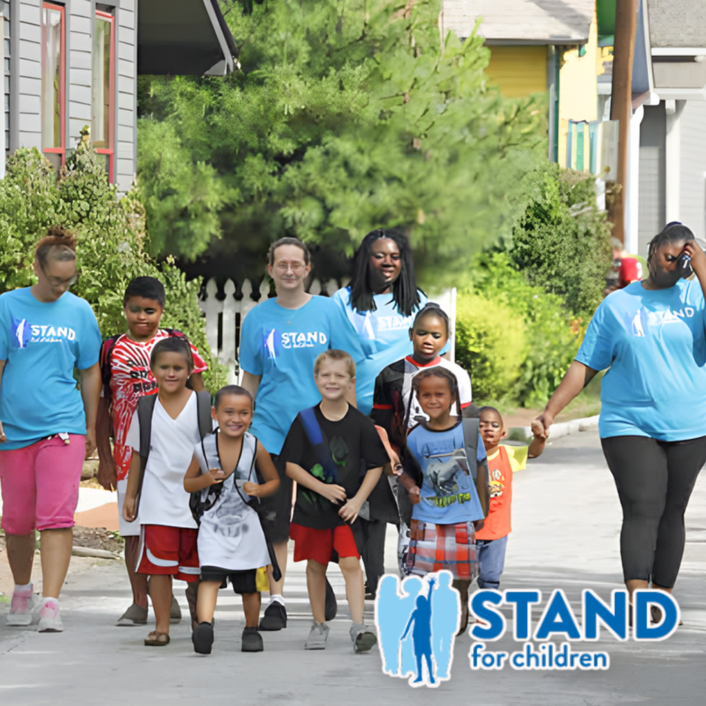 A group of adults and children walk together outdoors. The adults wear matching blue Stand for Children T-shirts, supporting youth empowerment. The children smile and walk hand in hand. The Stand for Children logo is in the lower right corner.