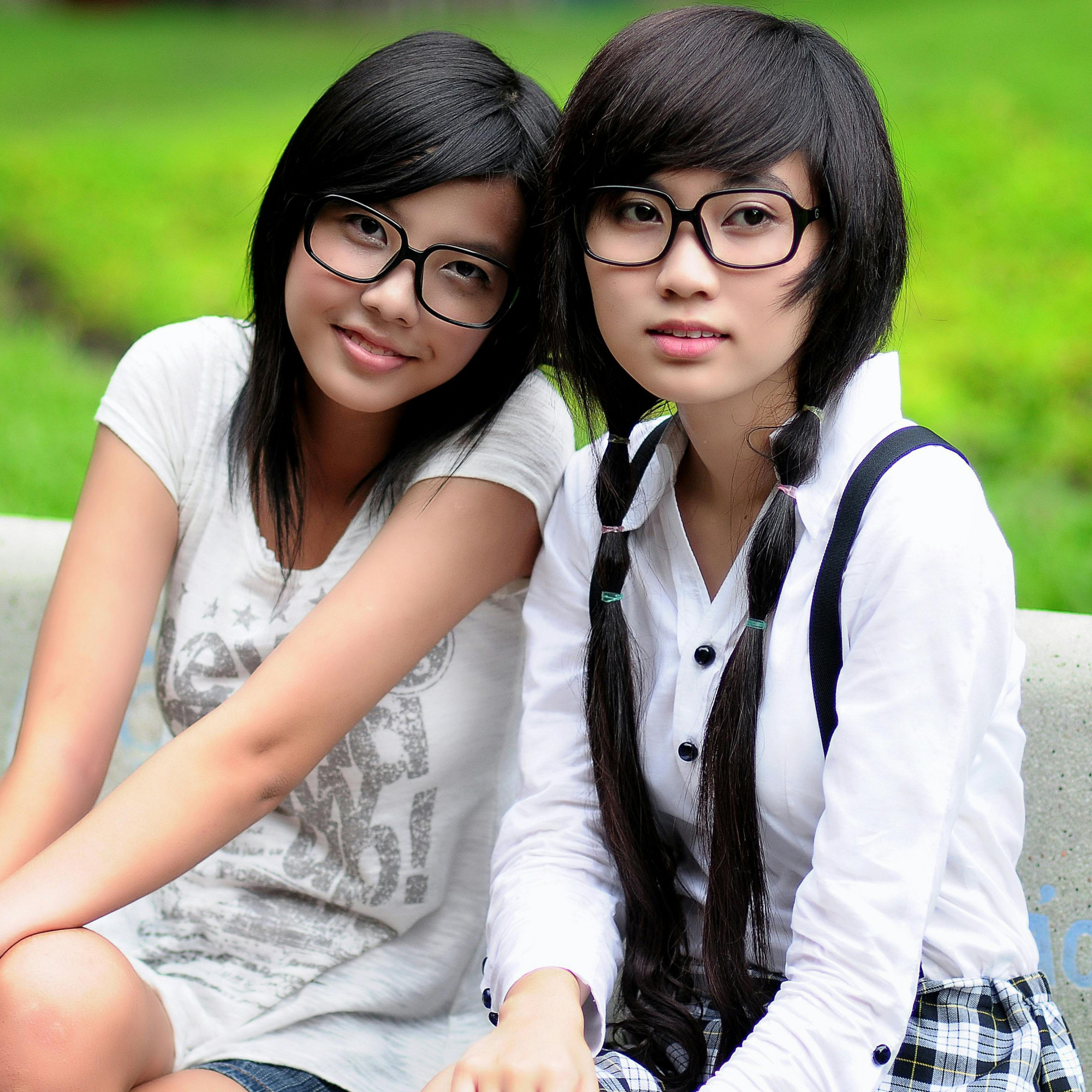 Two young women with glasses sit close together on a bench outdoors, smiling brightly. Perhaps discussing dual strategies for charitable services, they enjoy the greenery in the background—one in a white t-shirt, the other with suspenders and braided hair.