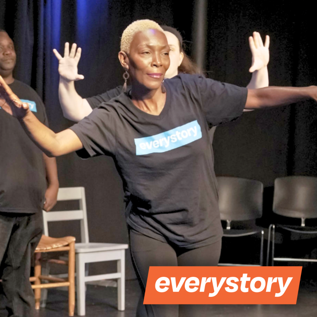 A woman with short blond hair, wearing a black everystory t-shirt, extends her arms while performing on stage, sharing stories as others stand behind her. The everystory logo is at the bottom right corner.
