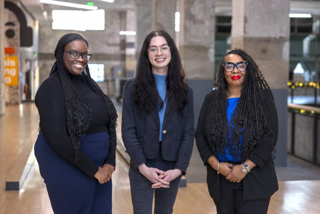 Three women stand side by side indoors, smiling at the camera. Dressed in business or smart-casual attire, they exude confidence against an industrial-style backdrop—suggesting a setting full of networking and job opportunities.