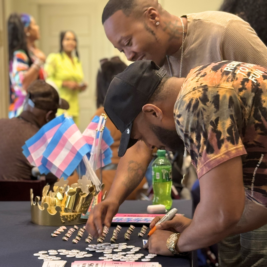 Two people lean over a table covered with buttons, markers, a crown prop, and mini transgender pride flags. Others are in the background, including someone in a bright yellow outfit. The setting appears to be an indoor event.