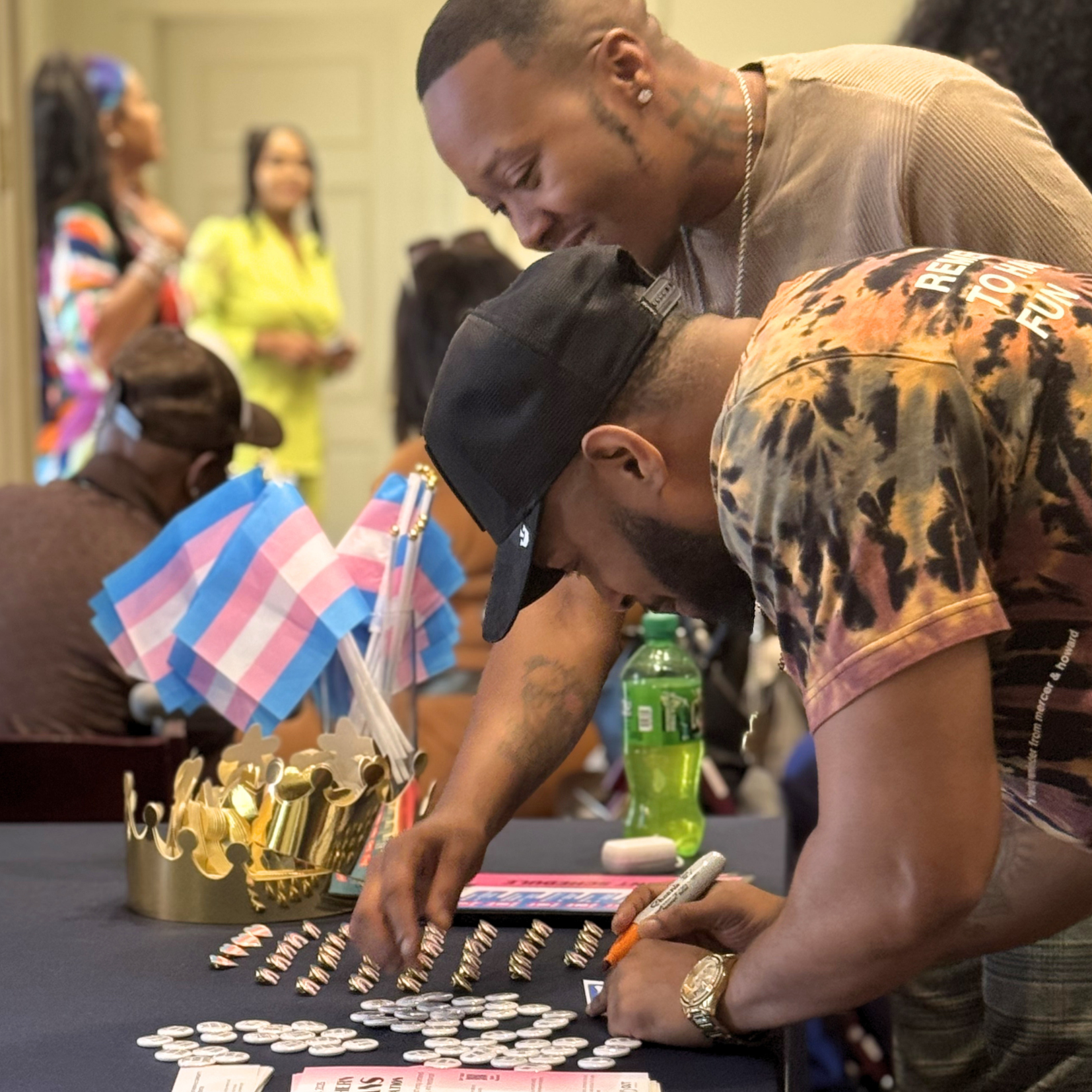 Two people lean over a table covered with buttons, markers, a crown prop, and mini transgender pride flags. Others are in the background, including someone in a bright yellow outfit. The setting appears to be an indoor event.