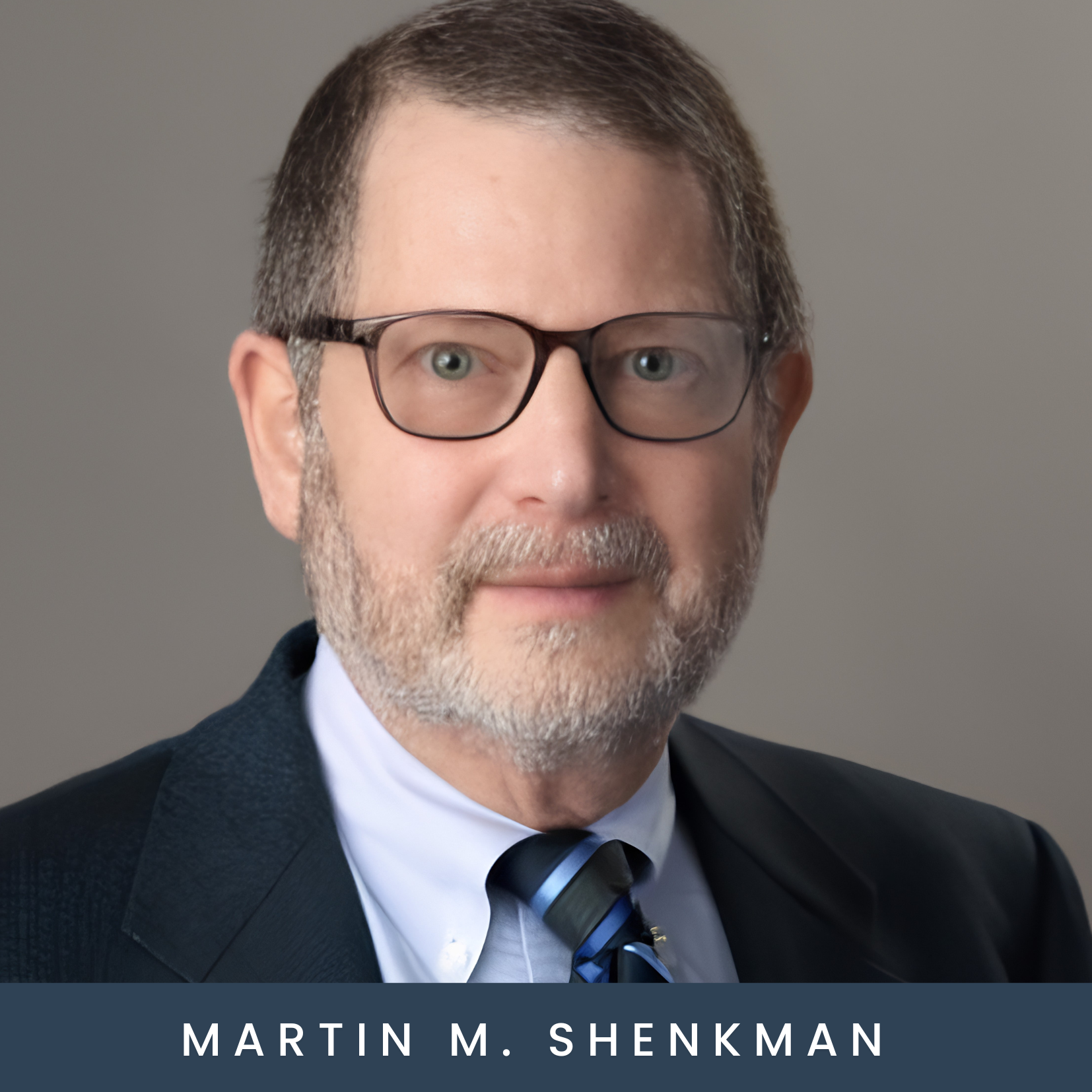 A man with short brown and gray hair, beard, and glasses, wearing a suit, light blue shirt, and patterned tie, poses in front of a plain gray background. Text below reads: Martin M. Shenkman.