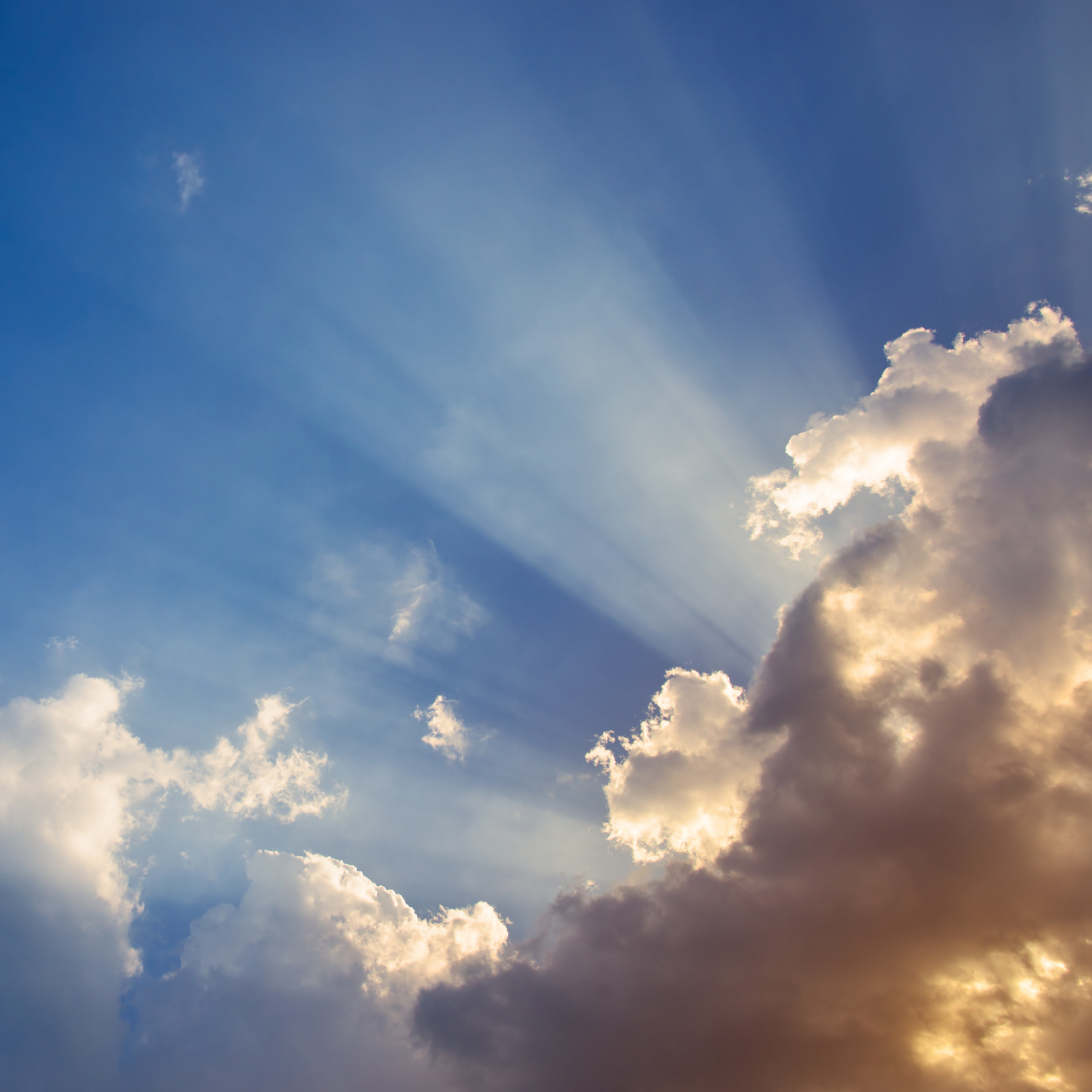 Sun rays shine through fluffy clouds in a blue sky, creating bright beams of light and a dramatic, peaceful scene. Some parts of the clouds are illuminated, while others are in shadow.