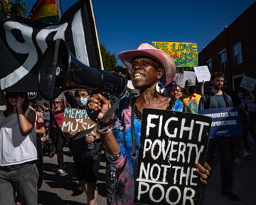 A person in a pink hat speaks into a megaphone at a protest, holding a sign that reads Fight poverty not the poor. Other people march behind, holding colorful signs and banners.