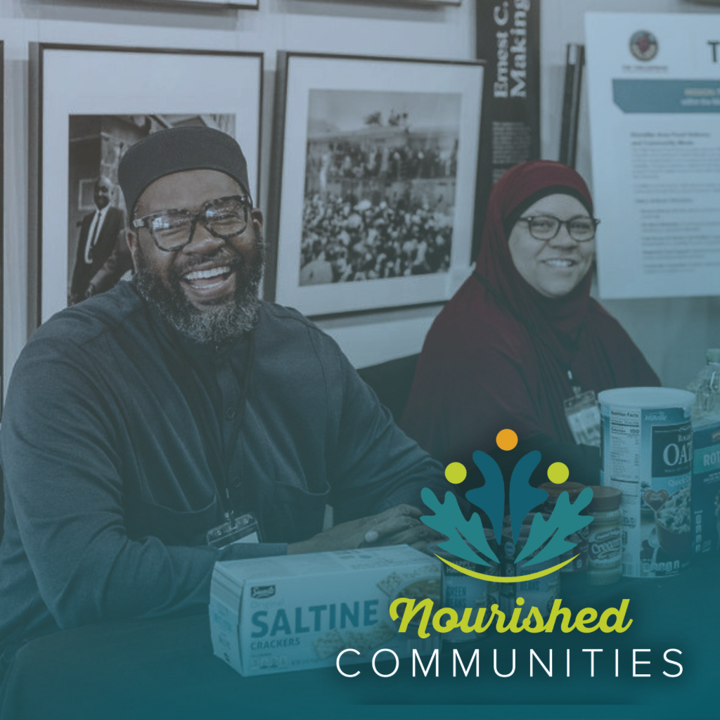 Two smiling people sit at a table with food items like saltine crackers and canned soup, showcasing community support. Behind them are framed photos and informational posters, with the Nourished Communities logo overlaid on the image.