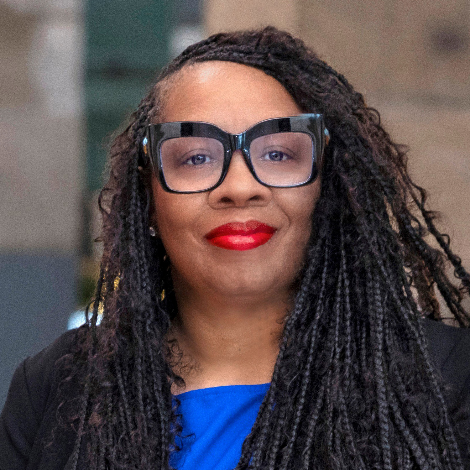 A woman with long braided hair, wearing oversized black glasses, bright red lipstick, a blue top, and a black blazer, stands in front of a blurred indoor background.