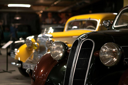 Close-up of the front of a classic black car with round headlights, parked inside a dimly lit garage alongside other vintage cars, including a yellow one in the background.
