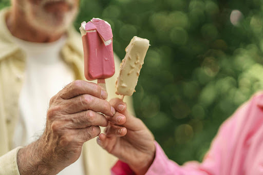 Two people holding ice cream bars, one pink and one white with nuts, about to toast them together. The background is blurred greenery, and only their hands and part of their torsos are visible.
