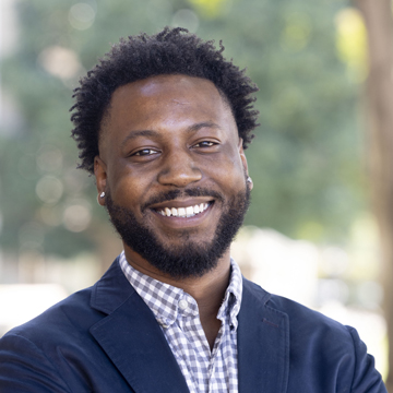 Smiling man, Adrian Williams, with short curly hair and a beard, wearing a blue blazer and checkered shirt, standing outdoors with blurred trees in the background.