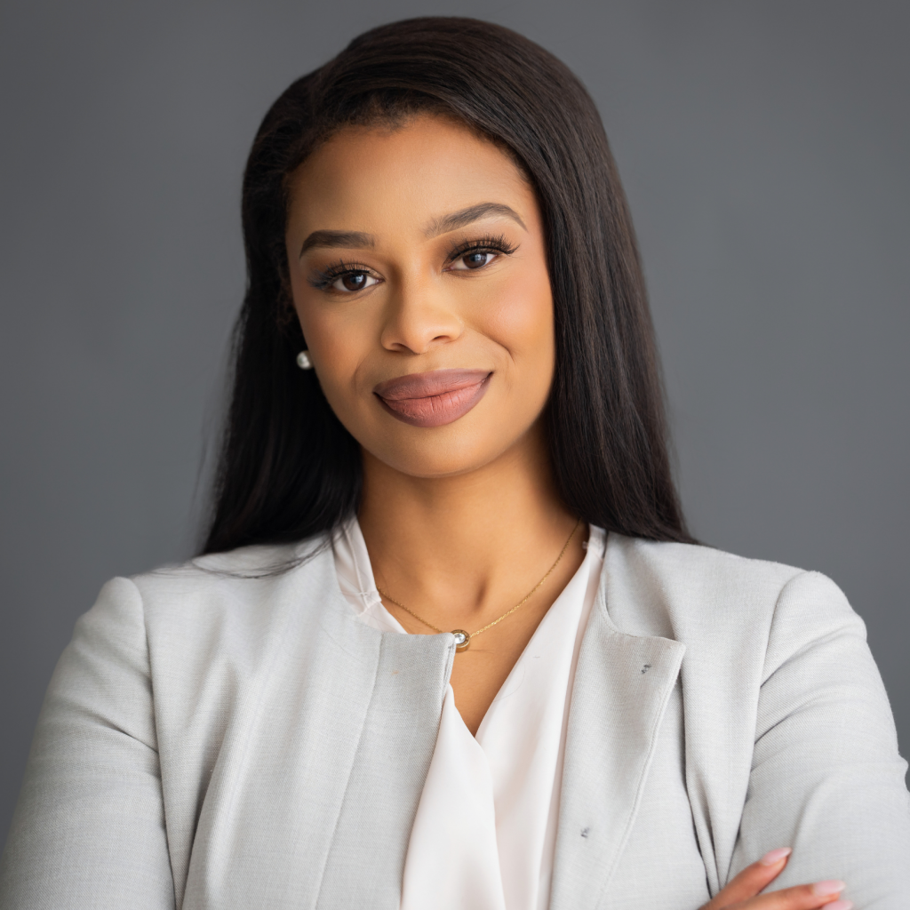 A professional headshot of a Black woman with long, straight dark hair, wearing a light grey blazer and a white blouse, smiling softly against a solid grey background.