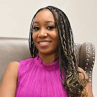 A woman with long braided hair, wearing a sleeveless magenta top, smiles while seated in a gray chair with silver studs. Chasity Grice exudes confidence and warmth in this poised moment.