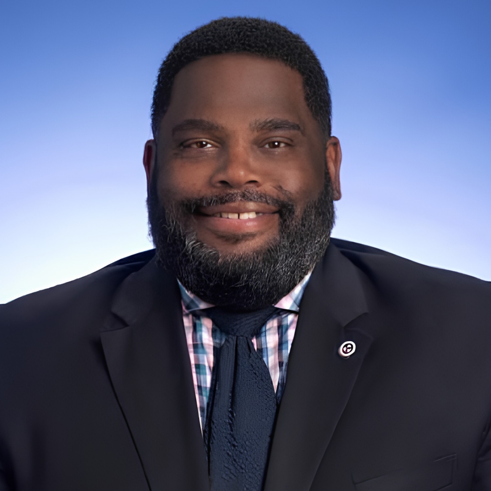 A man with a beard and short hair wearing a dark suit, plaid shirt, and navy tie, smiling in front of a blue background.