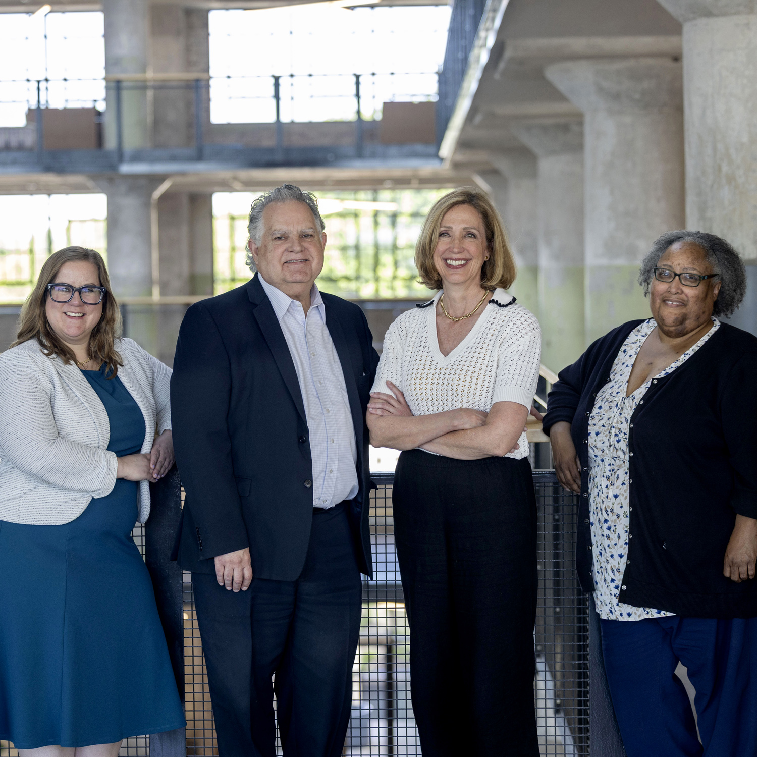 Four adults stand side by side indoors, smiling at the camera. Dressed in business or business-casual attire, they pose in front of a railing in a modern, industrial-style building—united in their commitment to building a legacy together.