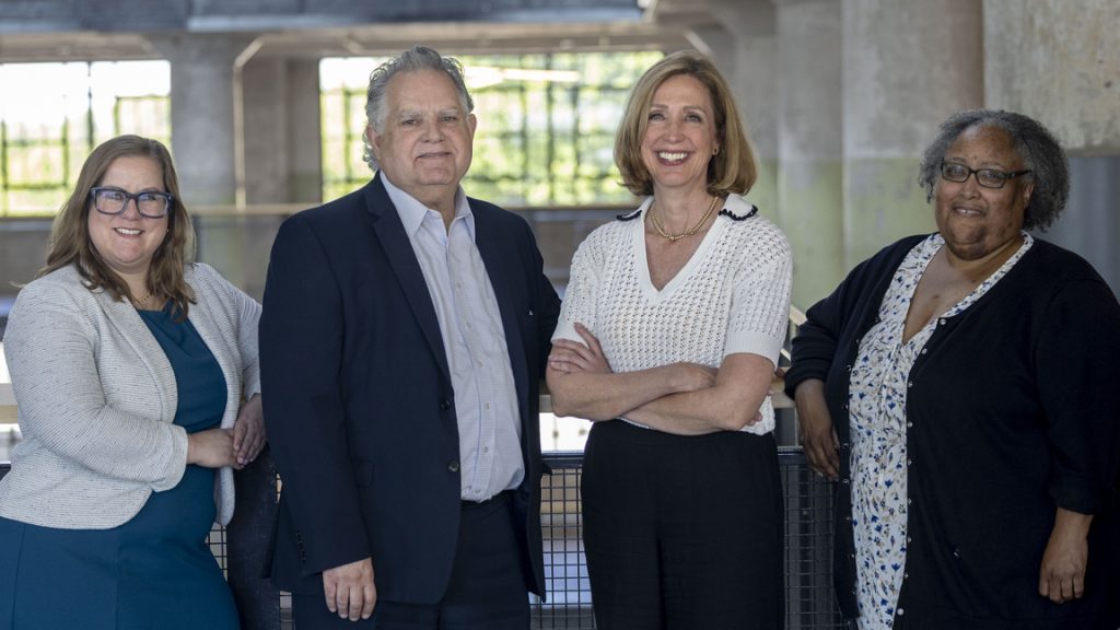 Four professionally dressed adults, two women on each side of a man and a woman, stand together indoors, smiling at the camera. The background features large windows and unfinished concrete walls—capturing a team dedicated to building a legacy.
