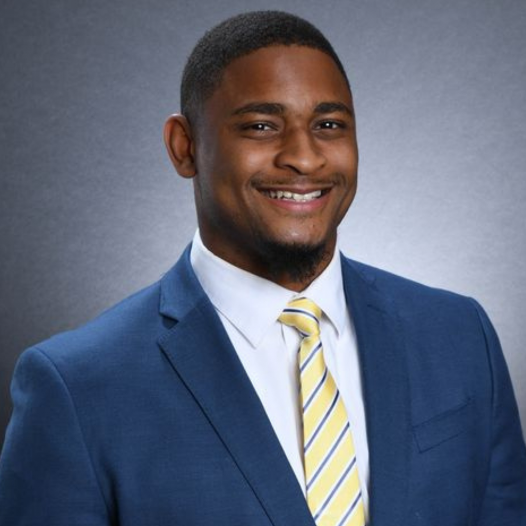 A man in a blue suit, white shirt, and yellow striped tie smiles at the camera against a plain gray background.