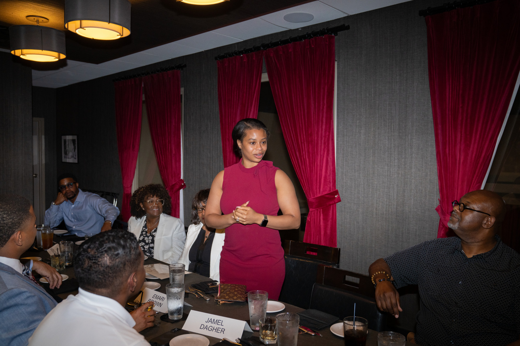 A woman in a red dress stands and speaks to a group at a dinner table in a dimly lit room with red curtains. Several people are seated, listening and smiling, with drinks and name cards on the table—an elegant gathering hosted by the Black Professional Advisor Alliance.