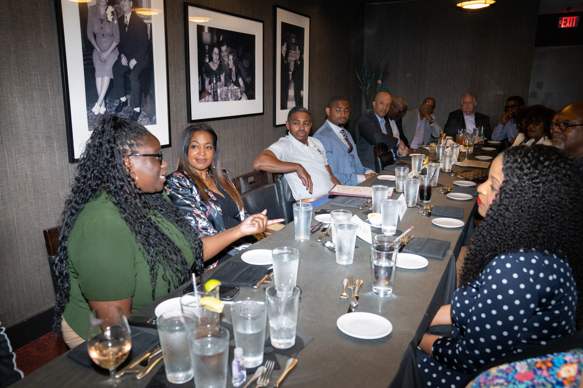 A group of people from the Black Professional Advisor Alliance sit around a long dining table in a restaurant, engaged in conversation. The table is set with glasses, plates, and silverware. Black and white photos hang on the wall behind them.