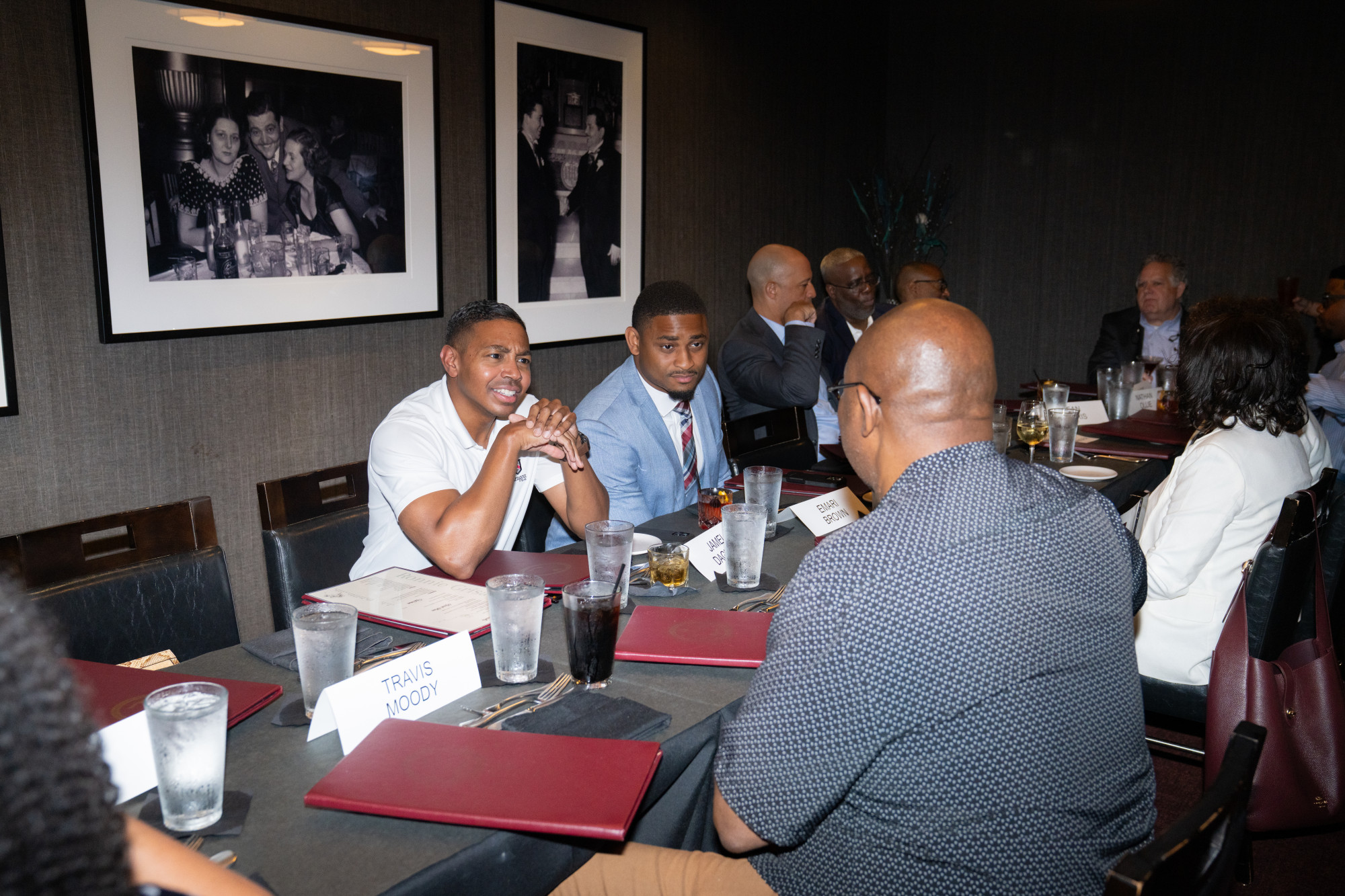 People in business attire, including several Black Professional Advisors, sit around a long table at a restaurant, talking and smiling. There are menus, drinks, and name cards on the table. Black-and-white photos decorate the dark walls, setting the scene for this Advisor Alliance gathering.