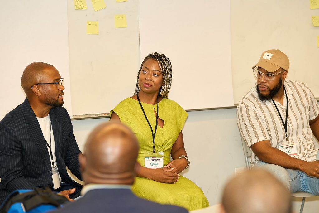 Three people sit and talk in a discussion panel. Two men and one woman, all wearing conference badges, face each other. Sticky notes are posted on the whiteboards in the background.