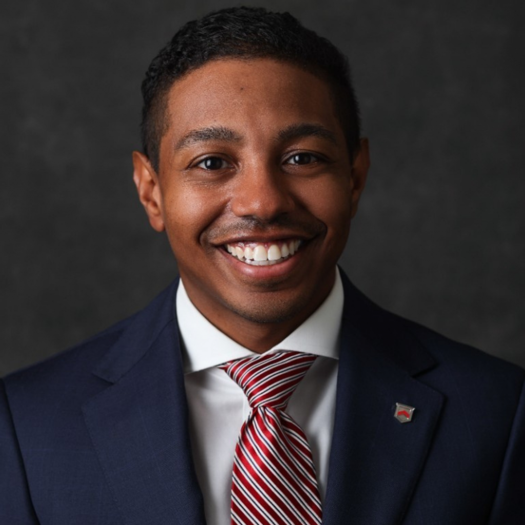 Smiling man in a dark suit, white shirt, and red striped tie poses in front of a dark background. He has short curly hair and a small pin on his lapel.