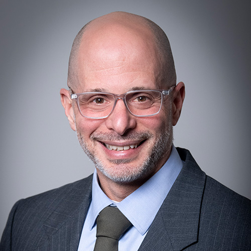 A smiling, bald man with a beard, identified as Jason Salomon, wears clear glasses, a gray suit, light blue shirt, and dark tie while posing in front of a plain gray background.