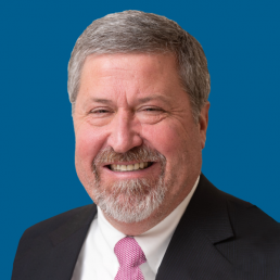Smiling middle-aged man, identified as Joel Kimbrough, with short gray hair, beard, and mustache, wearing a black suit, white shirt, and pink tie, posed in front of a solid blue background.